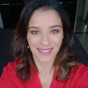 Daniele Schmitz, autora da tese indicada ao Prêmio CAPES de Tese 2025, com blusa laranja, cabelos castanhos ondulados e sorriso suave, em ambiente interno iluminado.