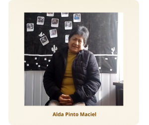 Mulher branca, de cabelos curtos e grisalhos. Usa uma blusa na cor mostarda sobreposta por um casaco preto, além de calça xadrez.