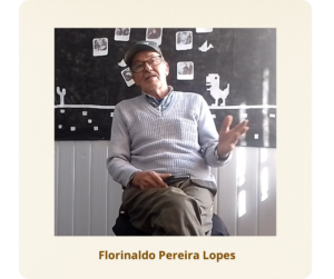 Homem branco, idoso. Veste um boné na cor grafite e óculos retangulares de armação marrom. Usa um suéter cinza sobre uma camisa xadrez, além de uma calça social marrom clara.